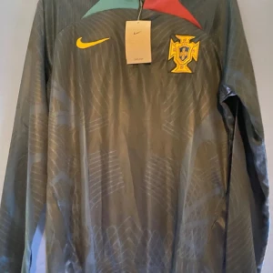 Portugal fotbollströja från Nike - Snygg Portugal fotbollströja från Nike med lång ärm och ett coolt mönster. Tröjan har en gul Nike-logga och ett emblem på bröstet. Perfekt för fotbollsfans som vill sticka ut på planen.