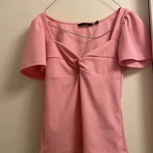 Rosa topp från Vero Moda - Säljer en söt rosa topp från Vero Moda med korta ärmar och en knutdetalj framtill. Perfekt för en somrig look. Toppen har en elegant och feminin design.