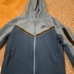 Blå fleecejacka från Nike junior/ barn storlek  - Säljer en blå fleecejacka från Nike med huva och dragkedja. Jackan har en mörkare blå detalj över bröstet och en ficka med dragkedja på ärmen. Perfekt för kyliga dagar. Små skador på tröjan se bilder. 