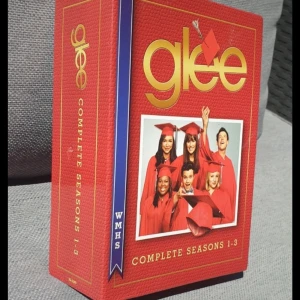 Glee Complete Seasons 1-3 DVD Box - Säljer en DVD-box med de kompletta säsongerna 1-3 av serien Glee. Boxen är röd med guldtext och innehåller flera skivor med alla avsnitt från de första tre säsongerna. Perfekt för Glee-fans som vill återuppleva serien från början.
