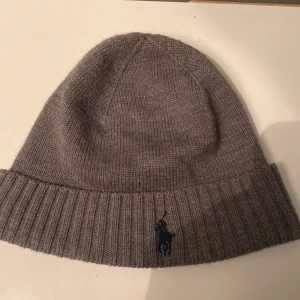 Grå mössa från Polo Ralph Lauren - Snygg grå mössa från Polo Ralph Lauren i mjuk merinoull. Den har en ribbad kant och en broderad logotyp i mörkblått framtill. Perfekt för kyliga dagar. Använd ett fåtal gånger. Bra skick