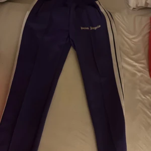 Palm Angels byxor - Snygga lila träningsbyxor från Palm Angels med vita och svarta ränder längs sidorna. Byxorna har en elastisk midja och sidofickor, perfekt för en sportig look. Helt nya,  Pris kan diskuteras.