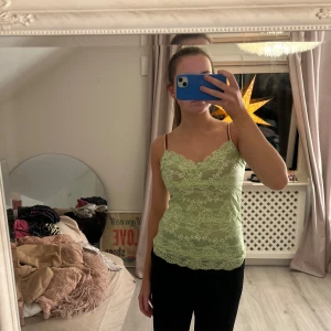 Grön spetslinne - Säljer ett snyggt grönt spetslinne med smala axelband och v-ringad design. Perfekt för en somrig look eller att bära under en kavaj. Linnet har ett vackert spetsmönster som ger en romantisk touch.