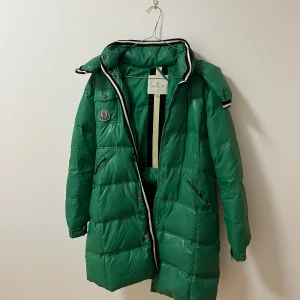 Moncler jacka - Säljer en snygg grön dunjacka från Moncler med huva och dragkedja. Jackan har långa ärmar och en stilren design med Moncler-logga på bröstet. Perfekt för kyliga dagar! (Pris kan diskuteras)