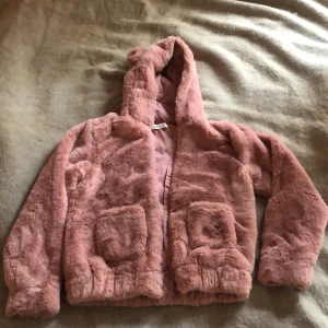 Rosa pälsjacka från H&M - Mysig rosa jacka från H&M med huva och två fickor framtill. Jackan är tillverkad i 100% polyester och har en fluffig look.