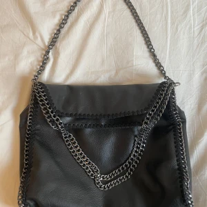 Svart axelväska med kedjedetaljer - Snygg svart axelväska i syntetmaterial med coola kedjedetaljer. Perfekt för att ge din outfit en edgy touch. Väskan har en stilren design och är rymlig nog för dina dagliga nödvändigheter.