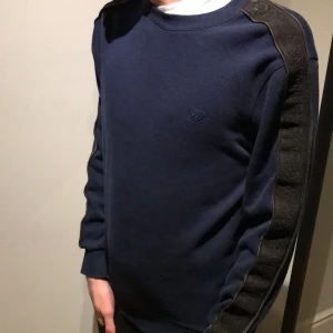 Mörkblå tröja från Emporio Armani - Snygg mörkblå tröja från Emporio Armani med svarta detaljer längs ärmarna. Tröjan har en diskret logga på bröstet och är perfekt för en stilren look. Äkta vara.