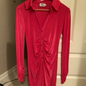 Rosa långärmad klänning - Snygg rosa klänning från NAKD i ett glansigt material. Klänningen har långa ärmar och en v-ringad design med knappar framtill. Perfekt för en färgglad look! Aldrig använd.