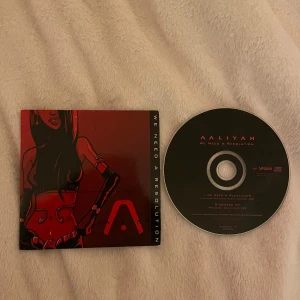 We Need a Resolution - Aaliyah - CD-singel av Aaliyah med titeln 'We Need a Resolution'. Omslaget är i rött och svart med en stiliserad illustration av en kvinna. Baksidan visar en närbild av ögon i samma färgtema. Perfekt för samlare av 2000-tals R&B-musik.
