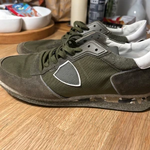 Olivgröna sneakers från Philippe Model - Snygga olivgröna sneakers från Philippe Model med en kombination av mocka och textil. Skorna har en klassisk snörning och en unik detalj på sidan. Perfekta för en avslappnad stil. Skorna är i bra skick förutom defekt på båda skorna vid sidan men påverkas it så mycket!