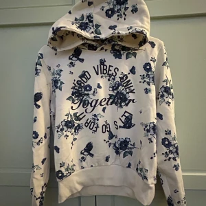 Vit blommig hoodie från KappAhl - Säljer en vit hoodie från KappAhl med blått blommönster och texten 'Good Vibes Only' på framsidan. Den har en mysig huva och ribbade muddar vid ärmslut och nederkant.