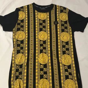 Svart och gul t-shirt med mönster från Versace - Snygg svart t-shirt från Versace med ett iögonfallande gult mönster av grekiska nycklar och medaljonger. Perfekt för dig som vill sticka ut med stil. Kortärmad och i normal passform. Ny pris 8800