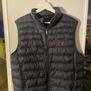 Ralph Lauren Väst - Mörkblå dunväst från Polo Ralph Lauren med dragkedja och broderad logga på bröstet. Perfekt för lager-på-lager under kyliga dagar. Västen har en stilren design och är lätt att matcha med olika outfits. Västen har en lös tråd på vänstra sidan men är inget som sticker ut och är väldigt lätt att laga. Nypris - 3500, Mitt pris - 950. 
