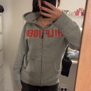 Grå zip hoodie från Tommy Hilfiger - Säljer en grå hoodie från Tommy Hilfiger med röd text på framsidan och en logga på ärmen. Den har en dragkedja och två fickor framtill. Anledningen till att jag säljer denna kofta är pågrund av att jag inte använder den lika ofta. Den var även ganska dyr och då den inte kommer till användning vill jag sälja den💕 (pris kan diskuteras)
