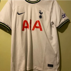 Vit Tottenham Hotspur fotbollströja - Säljer en vit Tottenham Hotspur fotbollströja från Nike med korta ärmar. Tröjan har klubbens logga och sponsortryck i rött på framsidan. Ärmsluten har en snygg detalj i mörkblått och gult. Perfekt för alla Spurs-fans! ⚽