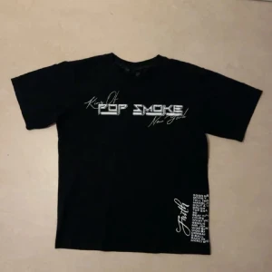 Vlone pop smoke tröja - Cool svart t-shirt med tryck av 'Pop Smoke' och 'Faith' i vitt. Stort 'V' med mönster på baksidan. Perfekt för streetwear-stilen. Riktigt snygg skriv vid fundering!
