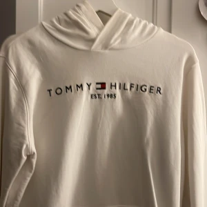 Vit hoodie från Tommy Hilfiger - Säljer en stilren vit hoodie från Tommy Hilfiger med logga och text 'EST. 1985' på bröstet. Tröjan har en klassisk känguruficka och en bekväm huva. Perfekt för en avslappnad look.