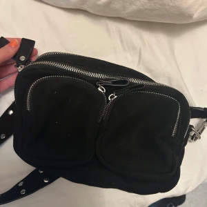 Svart axelväska med nitar - Snygg svart axelväska med två dragkedjeförsedda fickor framtill och en justerbar axelrem dekorerad med nitar. Kommer tyvärr inte till användning längre, mycket bra skick!