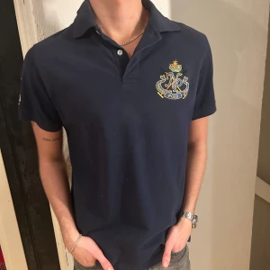 Mörkblå pikétröja  - Snygg mörkblå pikétröja från Polo med broderad logga på bröstet och siffran 4 på ärmen. Perfekt för en stilren look. Tröjan har en klassisk krage och knappar framtill. Storlek L/M