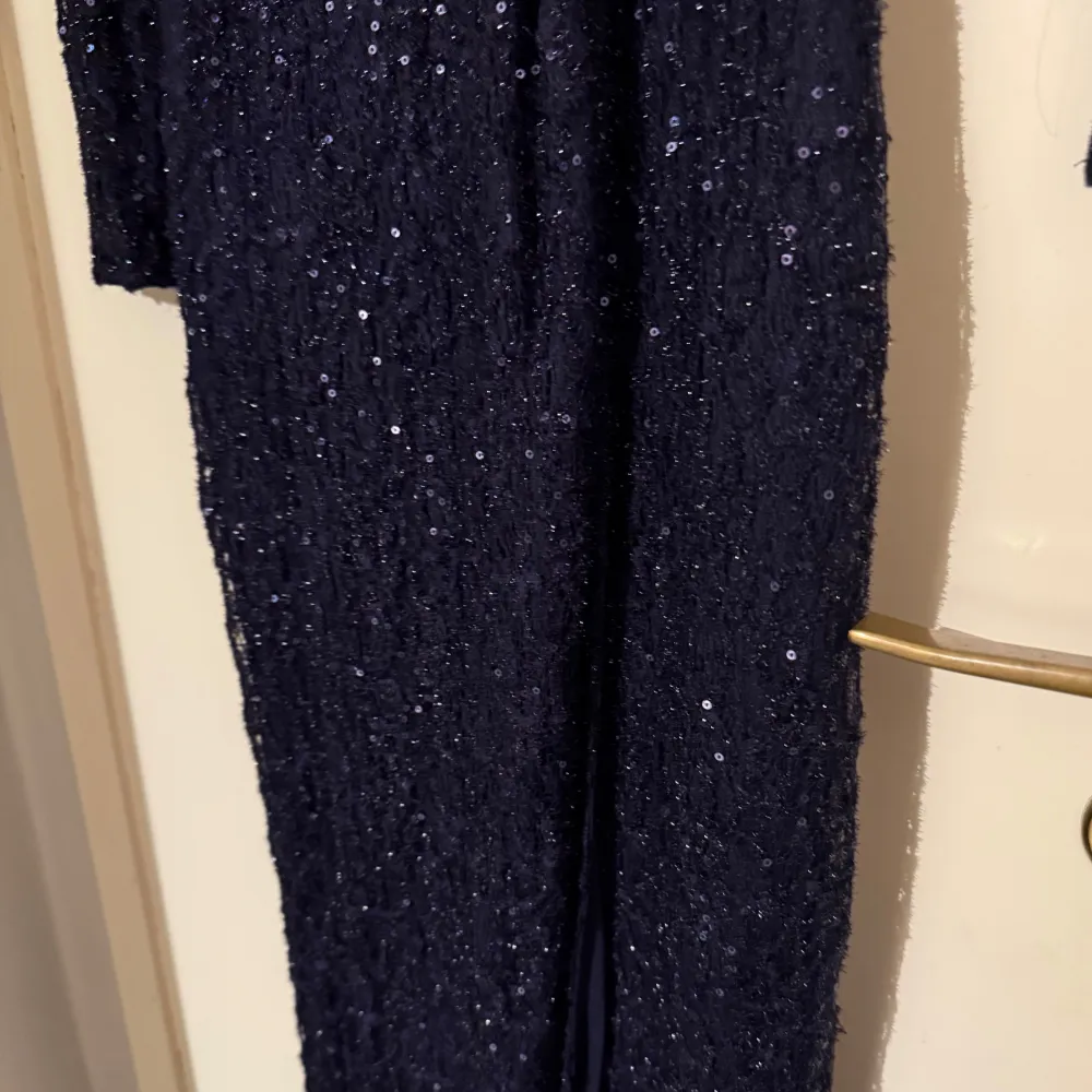 Elegant mörkblå klänning med glittriga detaljer. Den har långa ärmar och en knytning i midjan som ger en smickrande passform. Lång och delar sig framifrån. Perfekt för festliga tillfällen. Endast använt vid två tillfällen.. Mekot.