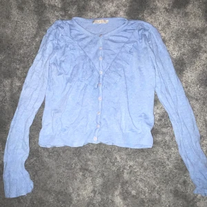 Ljusblå kofta med volangdetaljer - Söt ljusblå kofta med volangdetaljer längs knäppningen. Tröjan har långa ärmar och knappar framtill. Perfekt för en stilren och feminin look.