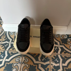 Svarta sneakers från Axel Arigato - Snygga svarta sneakers från Axel Arigato med vit sula och klassisk snörning. Perfekta för en stilren look. Skick 7-8/10, köpta för 2250kr! Kommer med Arigato boxen!