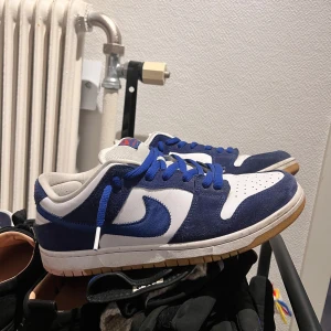 Nike SB Dunk la dodgers i blått och vitt - Snygga Nike SB Dunk sneakers i blått och vitt med klassisk snörning. Skorna har en vit bas med blå detaljer och en gummisula. Perfekta för en avslappnad stil. Bra skick, använd max 20 gånger.