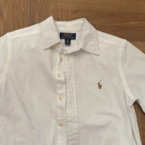 Vit kortärmad skjorta från Polo Ralph Lauren junior 8 år - Snygg vit kortärmad skjorta från Polo Ralph Lauren med klassisk krage och knappar framtill. Skjortan har en broderad logotyp på bröstet och är tillverkad i bomull för en bekväm passform.