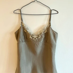 Top i 100% silke - Säljer ett användbart, beigeskimrande linne med spetsdetaljer. Linnet har smala axelband och en elegant, kvinnlig passform med rynk under bysten och en något lösare passform som faller snyggt. Perfekt under kavaj, till jeans och höga klackar eller sneakers. Eller till silkig kjol för att snabbt få en elegant look. 