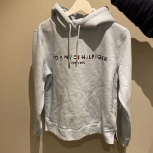 Ljusblå hoodie från Tommy Hilfiger - Säljer en stilren ljusblå hoodie från Tommy Hilfiger med broderad logga på bröstet. Tröjan har en klassisk huva med dragsko och långa ärmar. Perfekt för en avslappnad stil.