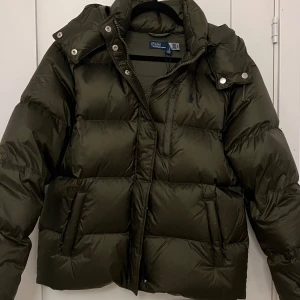 Ralph Lauren Dunjacka - Snygg grön dunjacka från Ralph Lauren med avtagbar huva och dragkedja framtill. Jackan har långa ärmar och praktiska fickor med knappar. Perfekt för kyliga dagar. Nypris 6695 kr 