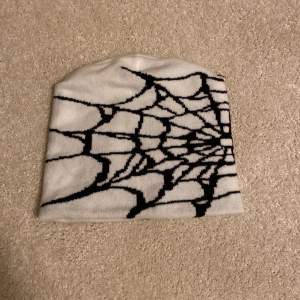 Snygg vit mössa med ett coolt svart spindelnätsmönster. Perfekt för att ge din outfit en unik touch. Passar bra till både höst och vinter.