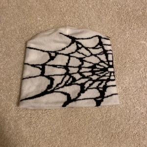 Vit mössa med spindelnätsmönster - Snygg vit mössa med ett coolt svart spindelnätsmönster. Perfekt för att ge din outfit en unik touch. Passar bra till både höst och vinter.