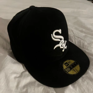 Fitted keps från New Era - Snygg svart keps från New Era med broderad vit logotyp på framsidan. Kepsen är en 59FIFTY-modell, vilket innebär en tight passform. Perfekt för sportiga tillfällen eller som en stilren accessoar.