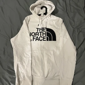 Vit hoodie från The North Face - Säljer en vit hoodie från The North Face med stor svart logga på framsidan. Den har en klassisk design med känguruficka och justerbar huva. Lite små fläckig men borde gå att tvätta bort i vanlig tvättmaskin