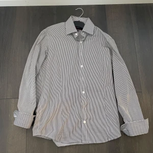 Randig skjorta - Snygg och stilren långärmad skjorta med smala ränder i svart och vitt ifrån dressman. Skjortan har klassisk krage och knappar framtill. Perfekt för en elegant look. Storlek M