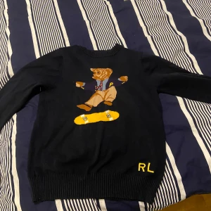 Svart tröja med björnmotiv från Ralph Lauren - Säljer en svart tröja från Ralph Lauren med ett coolt björnmotiv som åker skateboard. Tröjan har långa ärmar och RL-bokstäver broderade i gult vid nederkanten. Perfekt för en avslappnad stil! Hör av dig om du vill ha fler bilder. Priset är ej hugget i sten. 