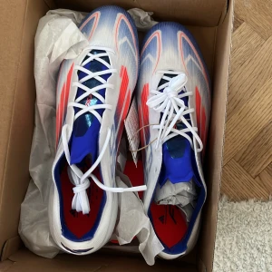 Adidas F50 fotbollsskor - Säljer ett par Adidas F50 fotbollsskor i vitt med blå och röda detaljer. Skorna har snörning och en modern design med dobbar för optimalt grepp på planen. Perfekta för fotbollsspelare som vill ha både stil och funktion.