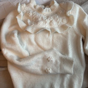 Vit stickad tröja med volangdetaljer - Säljer en söt vit stickad tröja med volangdetaljer runt halsen och blommiga broderier. Tröjan har långa ärmar och ribbade muddar. Perfekt för kyliga dagar!