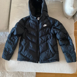 Svart dunjacka från Nike - Säljer en svart dunjacka från Nike med huva och dragkedja. Jackan har en klassisk design med Nike-logga på bröstet. Perfekt för kyligare dagar. Passar någon som är ca 145-160. Nypris ca 1500.