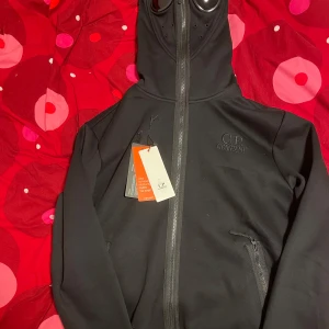 Svart fullzip hoodie från C.P. Company - Säljer en svart fullzip hoodie från C.P. Company med unik design. Jackan har en dragkedja framtill och en huva med inbyggda glasögon. Perfekt för en stilren look.