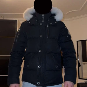 Svart dunjacka med pälsdetalj - Snygg svart dunjacka från Moose Knuckles med vit pälsdetalj på huvan. Jackan har dragkedja och knappar framtill samt flera fickor med dragkedjor. Perfekt för kalla vinterdagar. Köpt förra året för 14000kr.