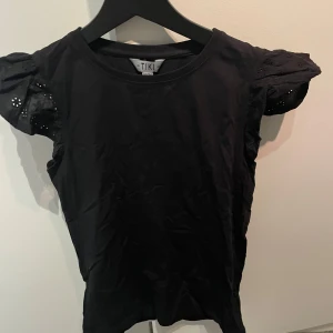 Svart topp från Tiki - Snygg svart topp från Tiki med puffiga kortärmar och spetsdetaljer. Perfekt för en stilren look.