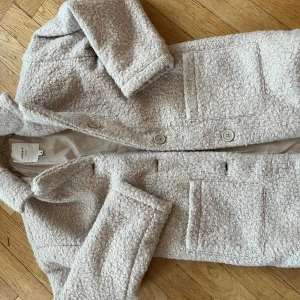 Beige kappa från Name It - Säljer en snygg beige kappa från Name It. Jackan har en texturerad yta med knappar framtill och två stora fickor. Perfekt för kyligare dagar och ger en stilren look.