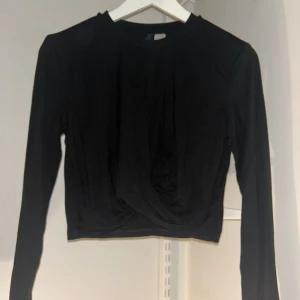 Svart långärmad topp - Snygg svart långärmad topp med en twistdetalj framtill. Perfekt för en stilren look och kan enkelt matchas med olika outfits. Toppen har en croppad passform som ger en modern touch.