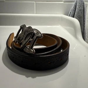 Svart bälte med dekorativt spänne - Snyggt svart Dsquared bälte med ett stort, dekorativt spänne i silverfärg. Bältet har ett detaljerat mönster ingraverat längs hela längden, vilket ger det en unik look. Perfekt för att ge din outfit en extra touch.