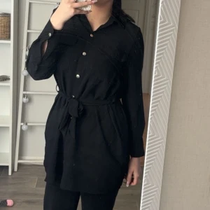 Svart skjortklänning med knappar - Snygg svart skjortklänning med knappar framtill och knytskärp i midjan. Klänningen har långa ärmar och en klassisk krage, perfekt för en stilren look.