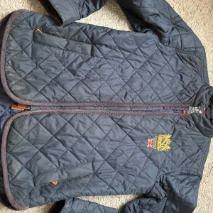 Mörkblå quiltad jacka från Morris - Snygg mörkblå quiltad jacka från Morris med broderad logga och brittisk flagga på bröstet. Jackan har dragkedja och långa ärmar med knappar vid ärmsluten. Perfekt för en stilren look.