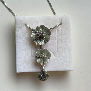 Silverfärgat halsband med blommor från Pilgrim - Elegant silverfärgat halsband från Pilgrim med tre vackra blommor i olika storlekar. Kedjan är tunn och justerbar med en liten logotypbricka vid låset. Knappt använd.