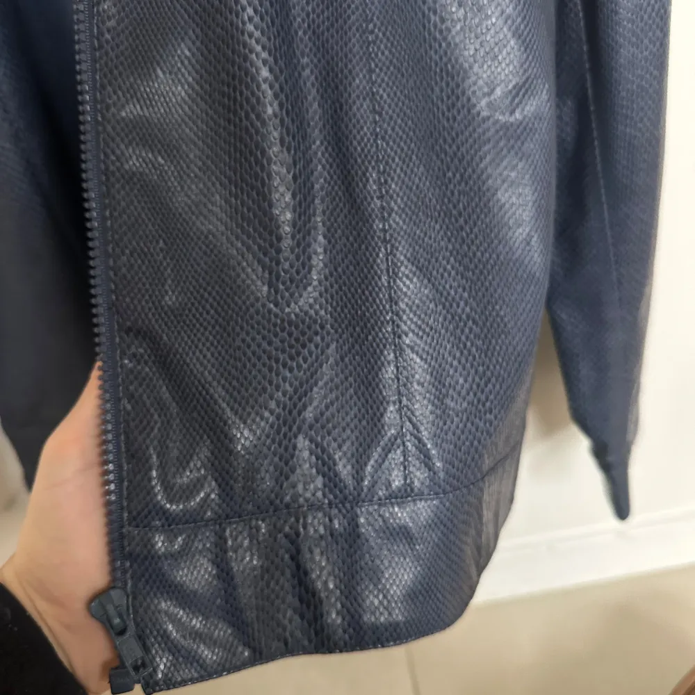 Snygg blå skinnjacka med texturerad yta och dragkedja framtill. Jackan har en stilren design med långa ärmar och en hög krage med knappdetalj. . Takit.